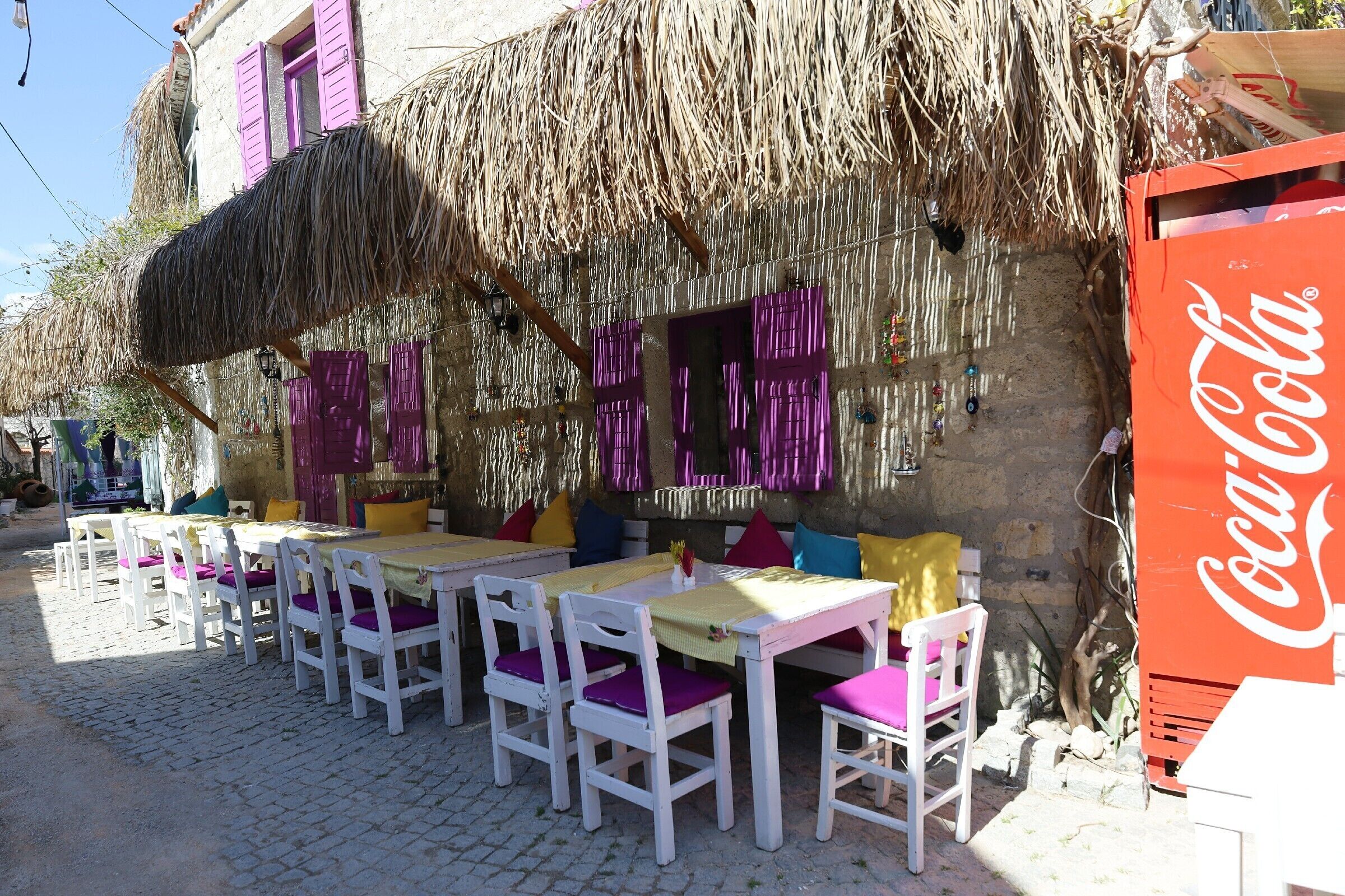 An outdoor seating area with white tables and purple chairs, a thatched roof, and a Coca-Cola refrigerator.