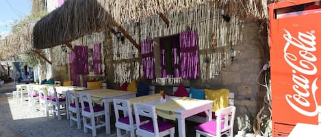 An outdoor seating area with white tables and purple chairs, a thatched roof, and a Coca-Cola refrigerator.