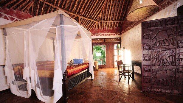 Habitación con cama bajo una mosquitera, techo tejido, puerta tallada y vista a la vegetación exterior.