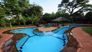 Una piscina con un diseño curvo singular, rodeada de exuberante vegetación y una estructura con techo de paja.