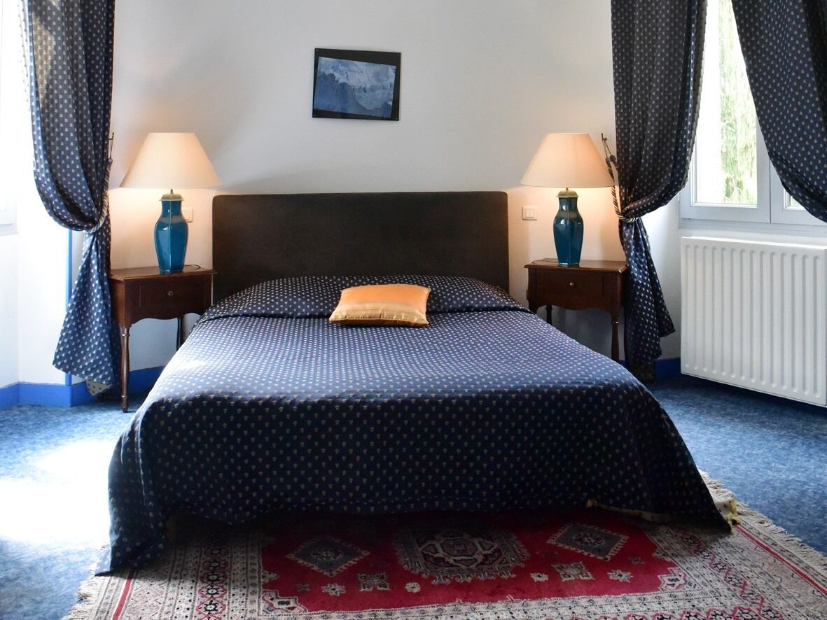 Een net opgemaakt bed met een bedsprei met een patroon en een enkel kussen, geflankeerd door twee nachtkastjes met lampen, een ingelijst schilderij aan de muur, en een raam met gordijnen.