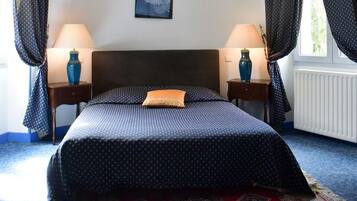 Een net opgemaakt bed met een bedsprei met een patroon en een enkel kussen, geflankeerd door twee nachtkastjes met lampen, een ingelijst schilderij aan de muur, en een raam met gordijnen.