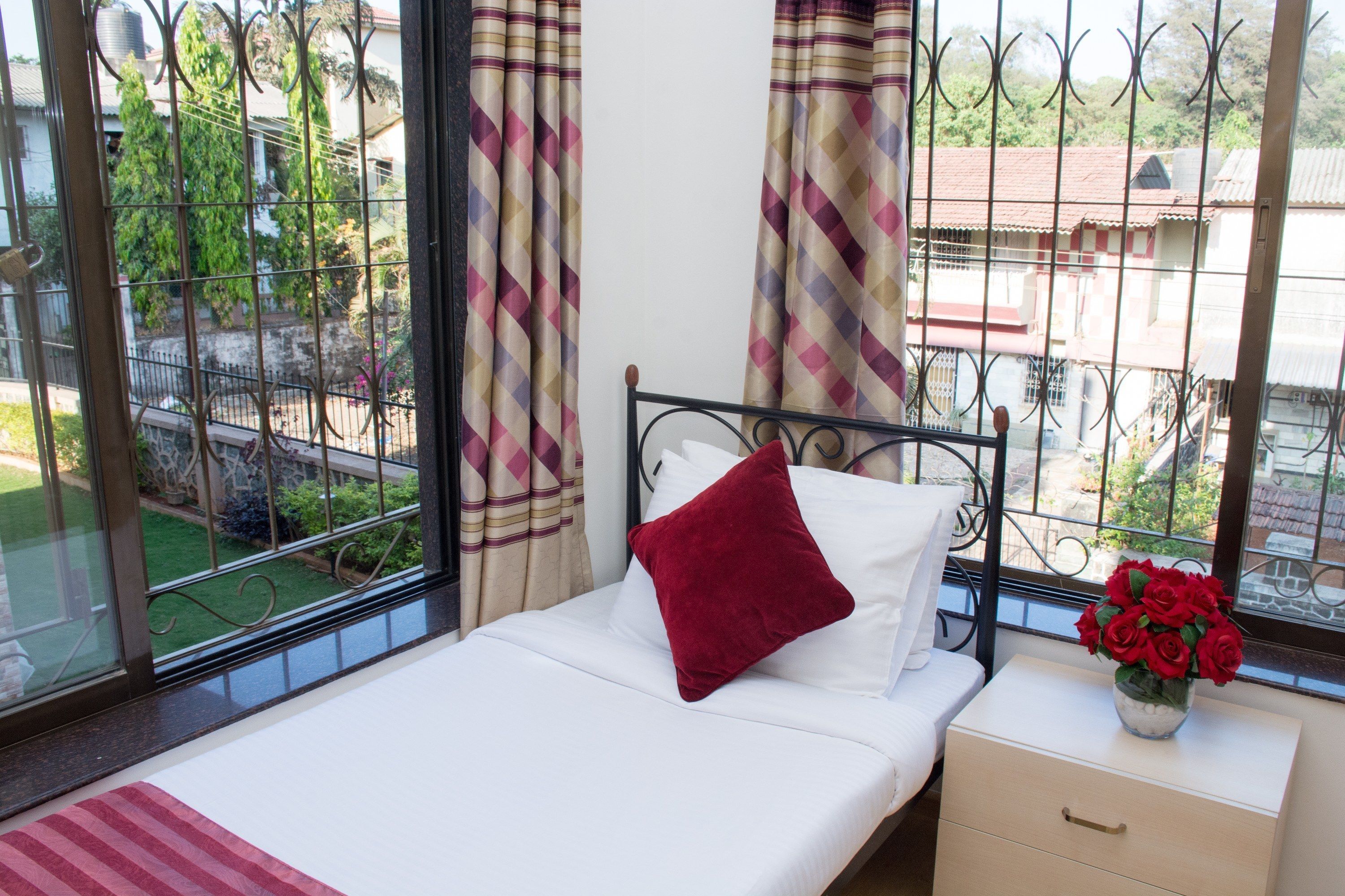 Ein Bett mit weißer Bettwäsche und einem roten Kissen, ein Nachttisch mit einer Vase voller roter Rosen und ein Blick auf einen Garten und Gebäude durch ein Fenster mit Metallgitter.