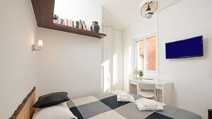 Un dormitorio con cama, escritorio, televisor y estante con libros y un florero.