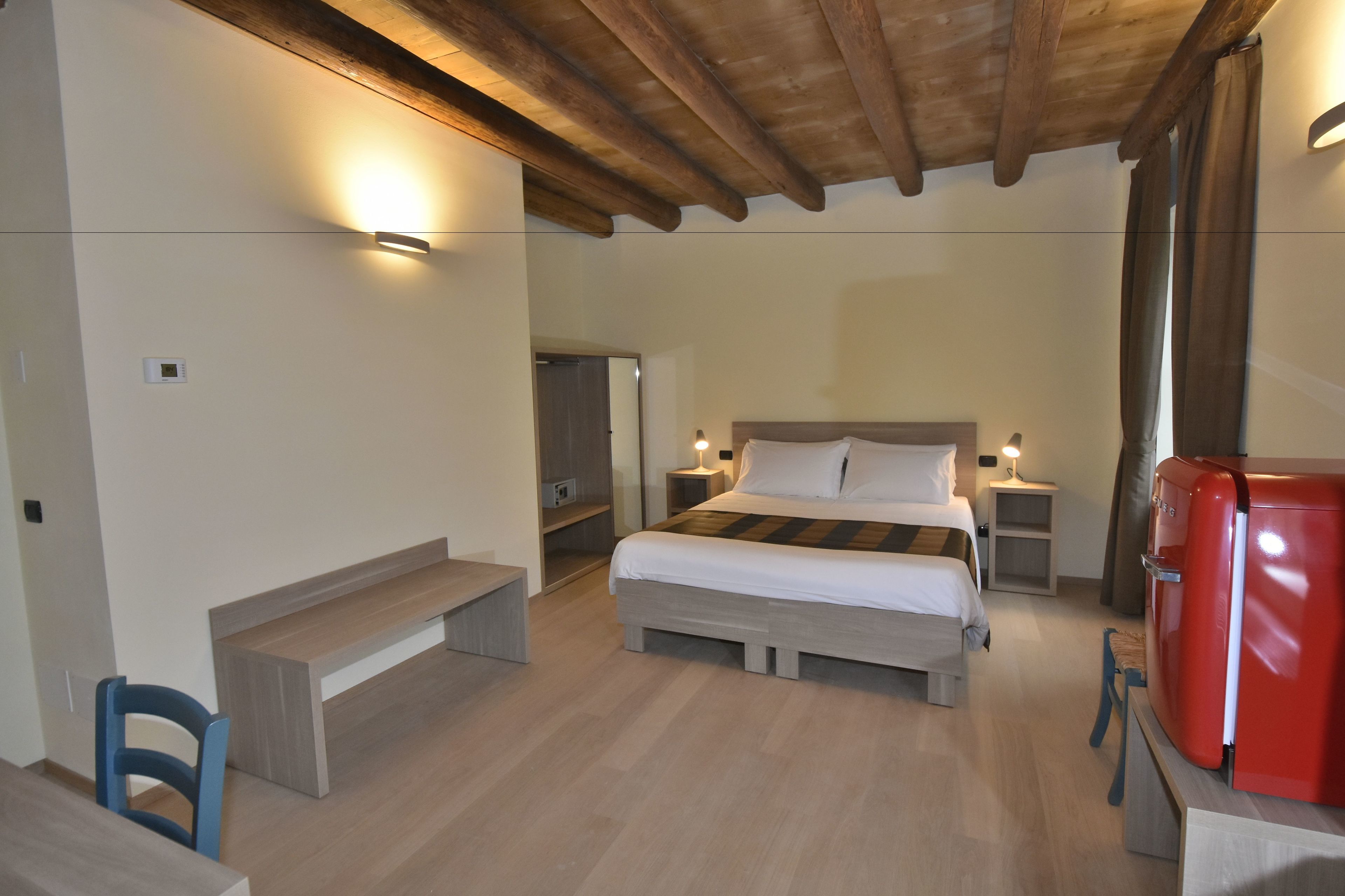 A bedroom with a bed, bedside tables, a chair, a small table, a red appliance, and a wooden ceiling.