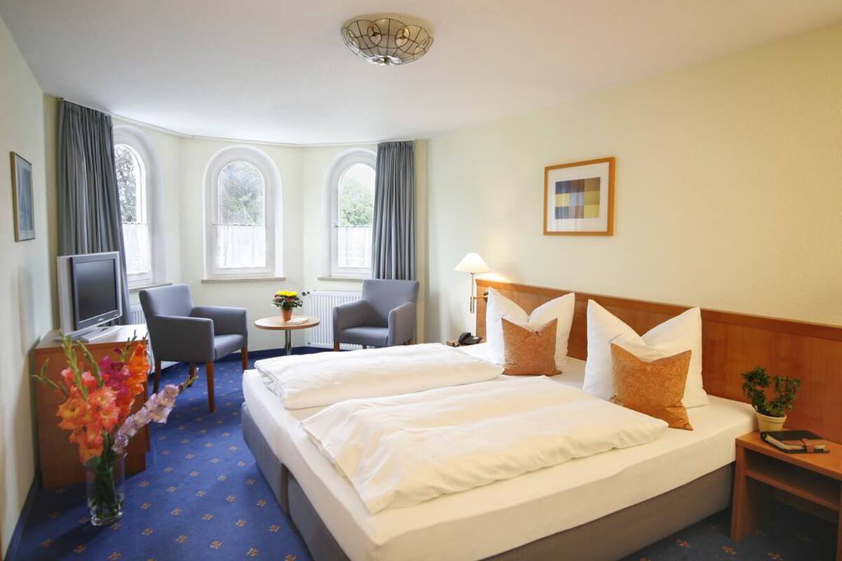 A hotel room with a large bed, two chairs, a small table, a TV, and a window with curtains.