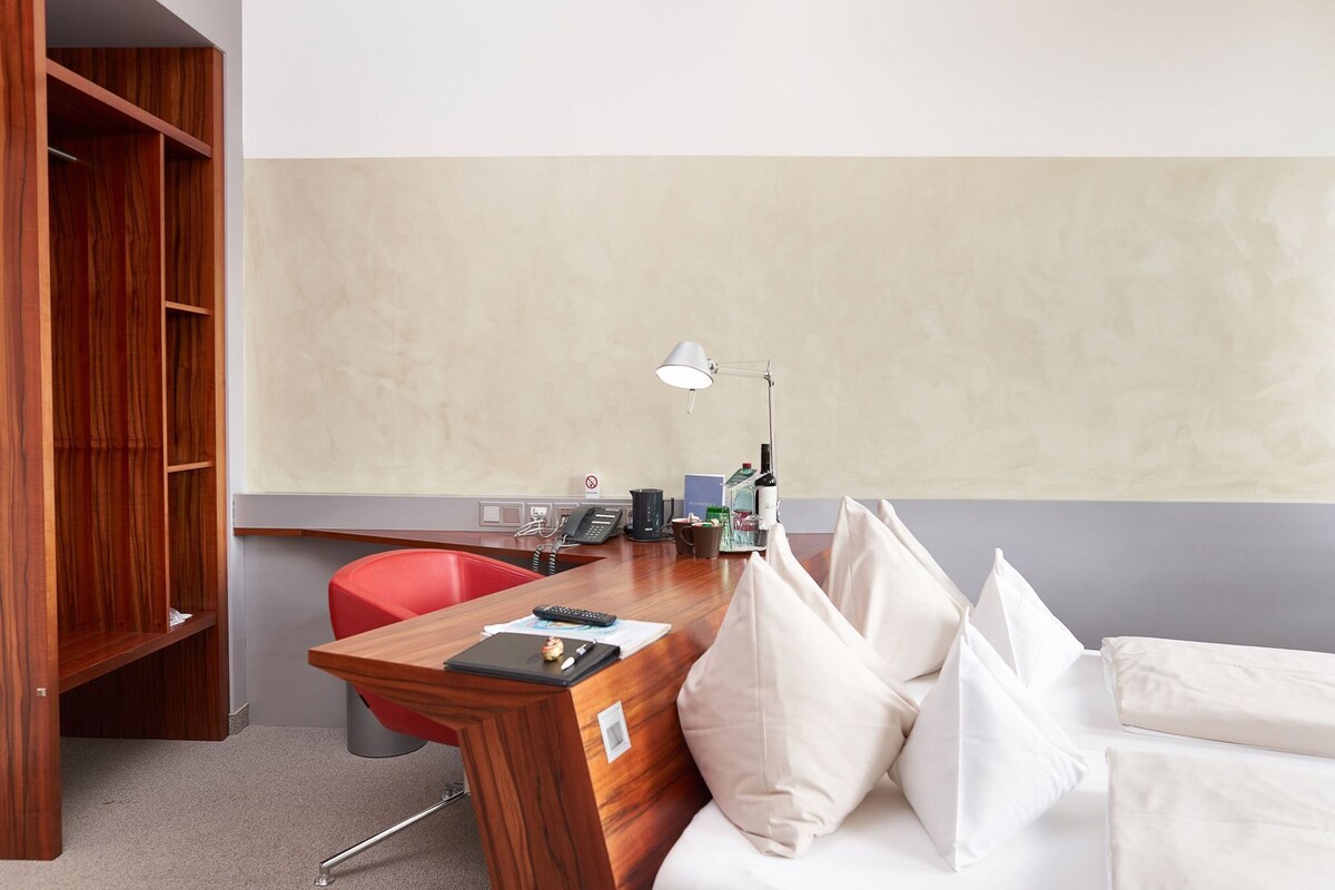 Habitación de hotel con un escritorio de madera, una silla roja, una cama blanca, una lámpara de escritorio, un teléfono y un minibar.