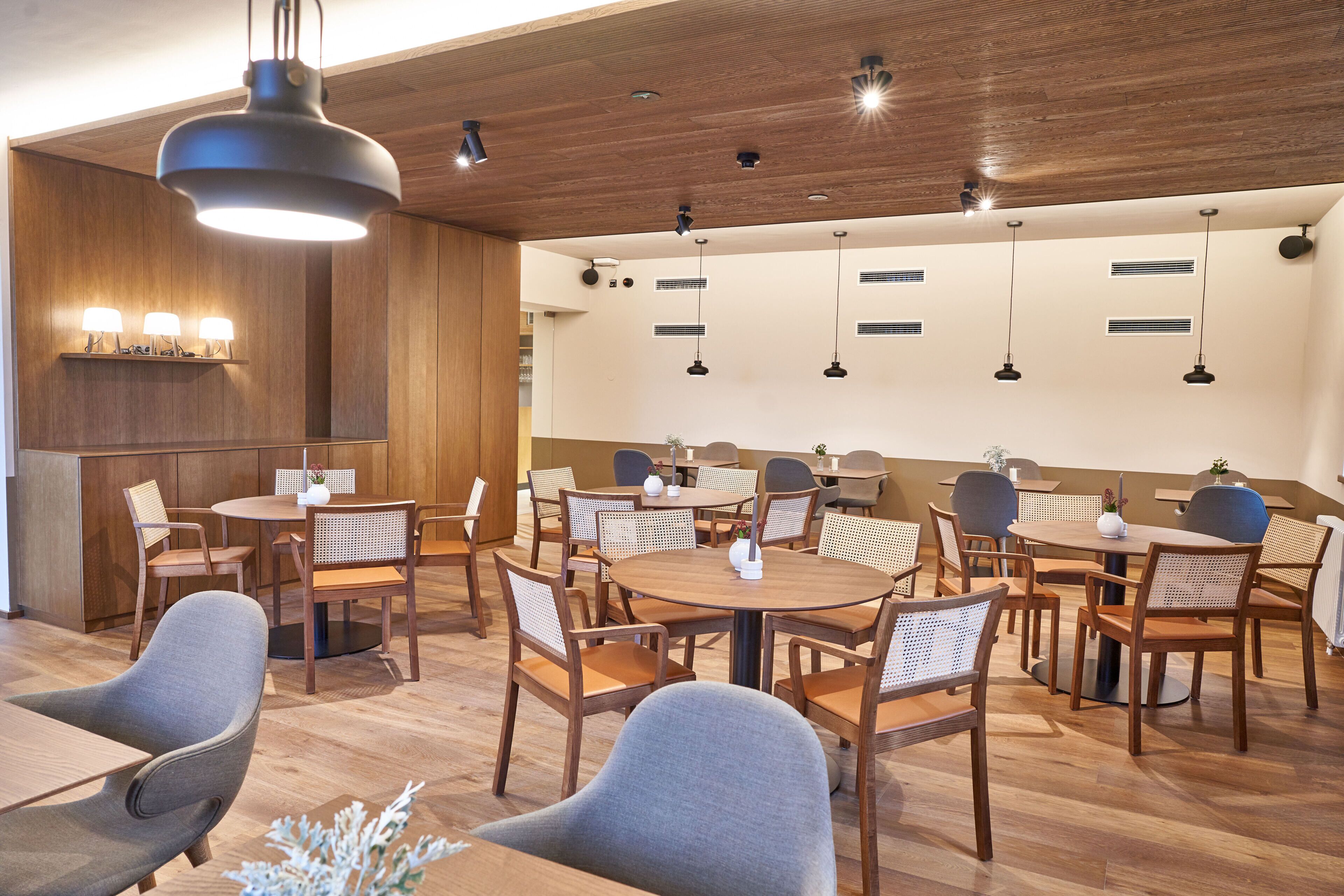 A modern dining area with wooden tables and chairs, pendant lighting, and a wooden ceiling.