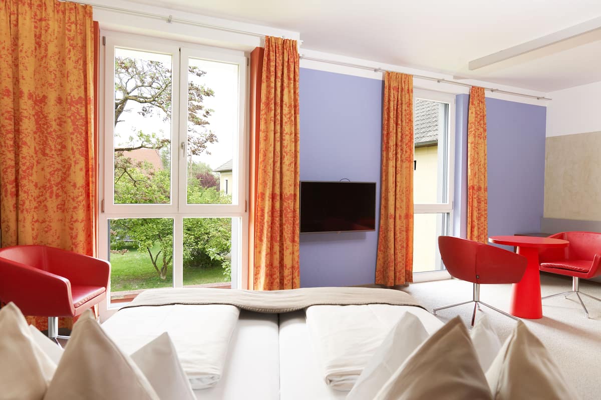 A hotel room with a bed, a TV mounted on the wall, a window with curtains, and red chairs.