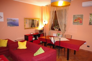 Un comedor con una mesa preparada para dos, un sofá rojo con almohadones amarillos, un espejo y un letrero de pizza en la pared.