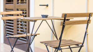 Una taula de fusta petita i dues cadires amb estructura metàl·lica, col·locades davant d'una porta amb mànec i una finestra amb persianes.