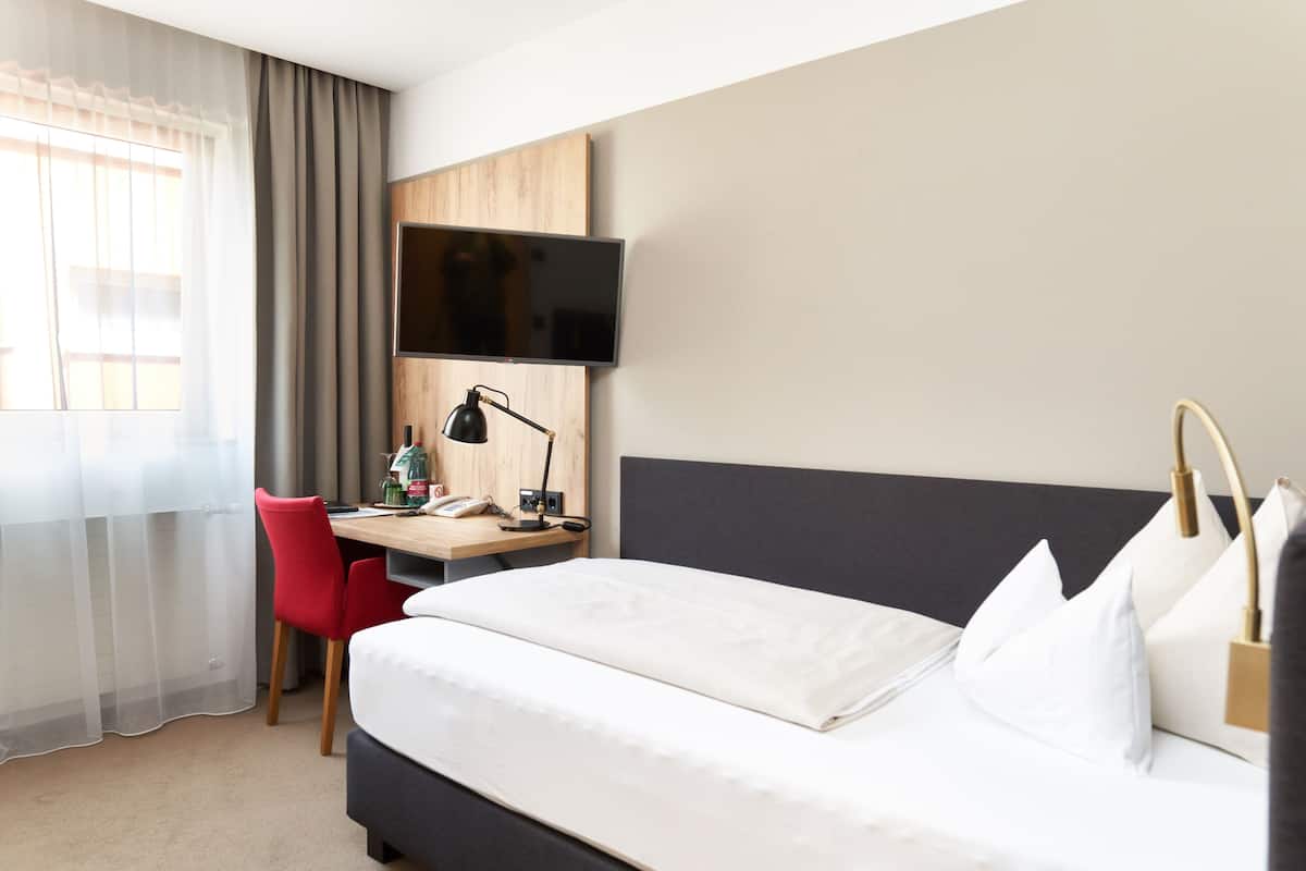 A hotel room with a bed, a desk with a computer, a chair, a TV mounted on the wall, and a window with curtains.