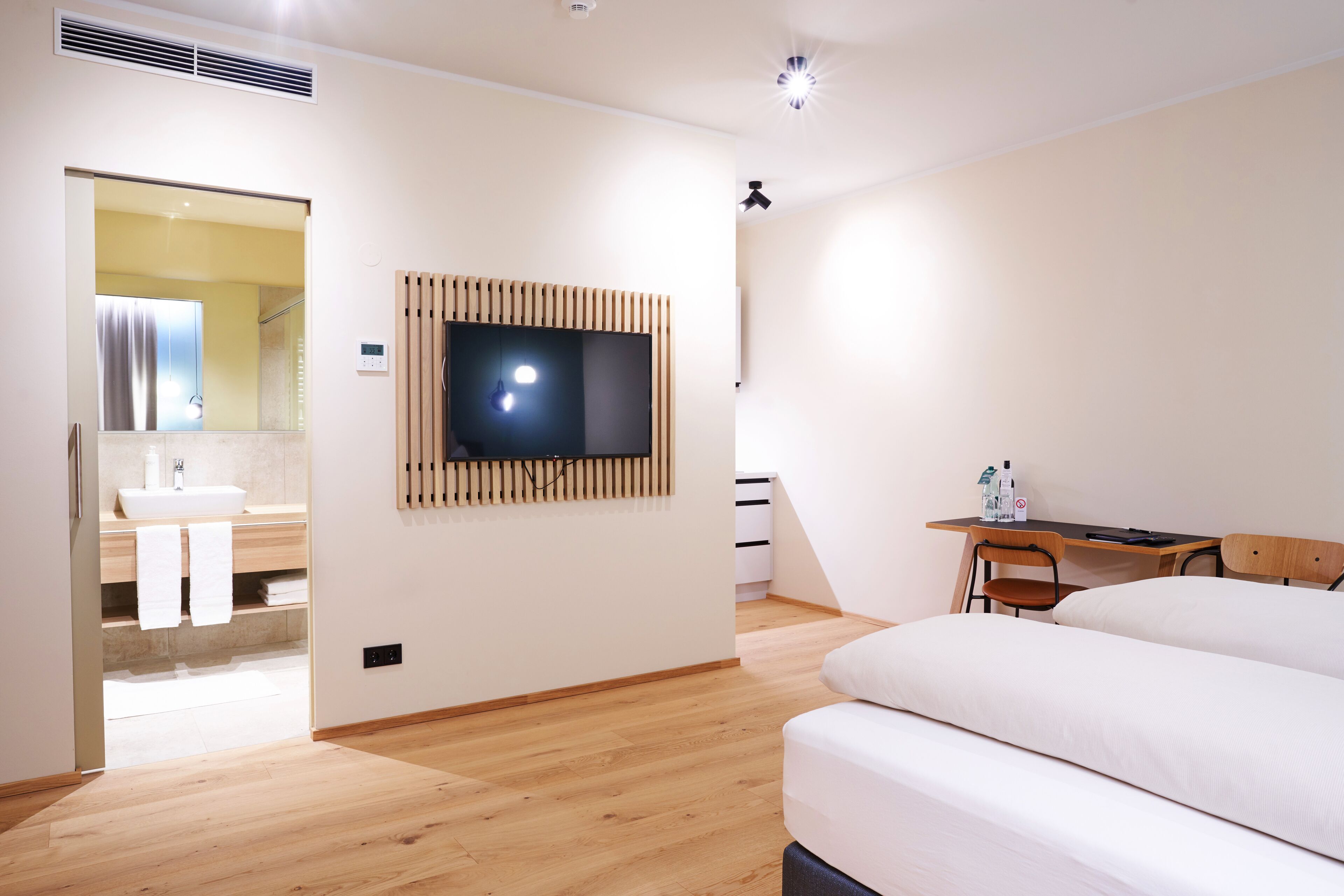 Una habitación de hotel moderna con suelo de madera, una cama, un escritorio con una silla, un televisor montado en la pared y un baño con lavabo y espejo.