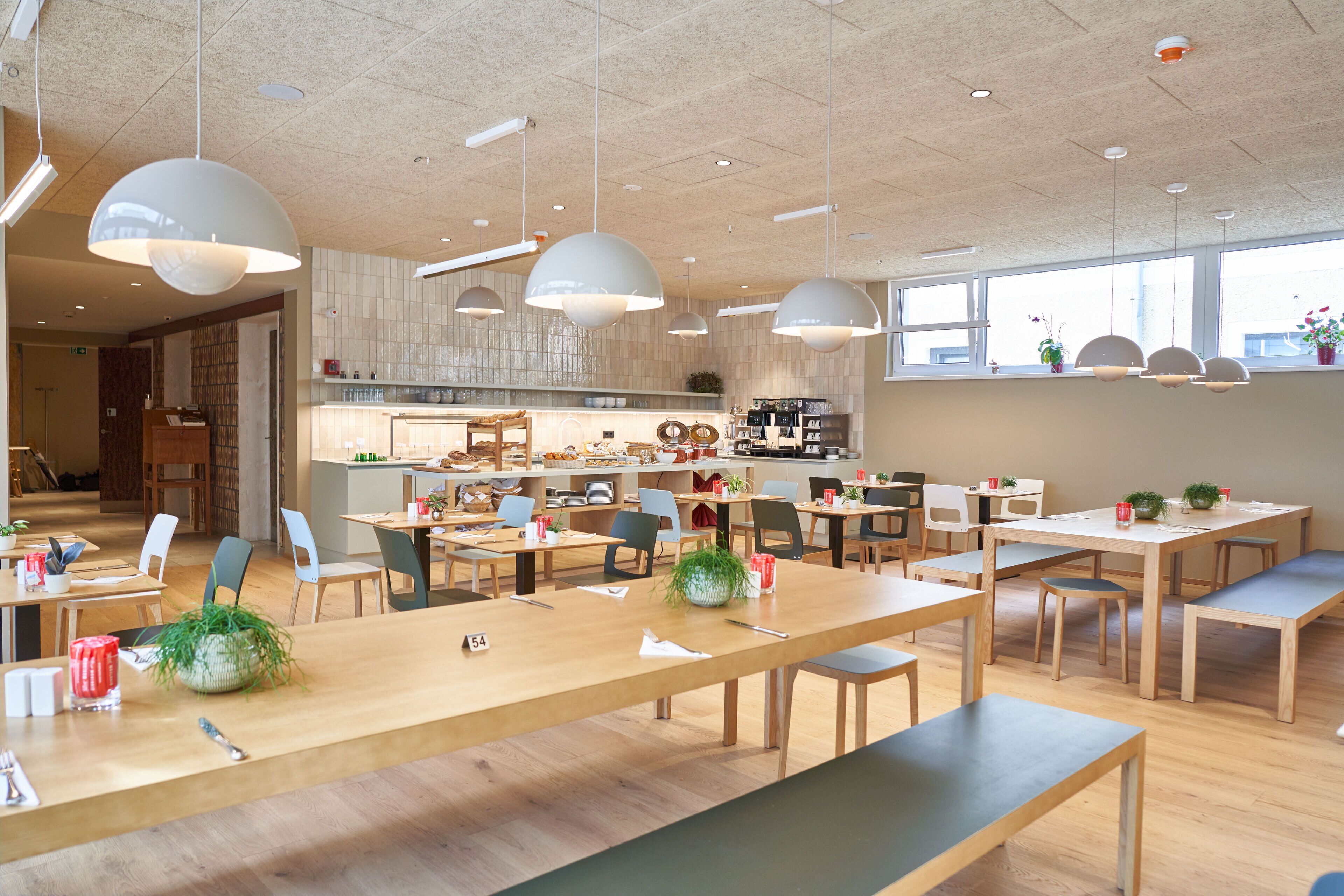 A spacious dining area with multiple tables and chairs, a kitchen in the background, and large windows allowing natural light.