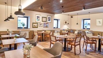 A spacious dining area with wooden tables and chairs, framed pictures on the walls, and pendant lighting.