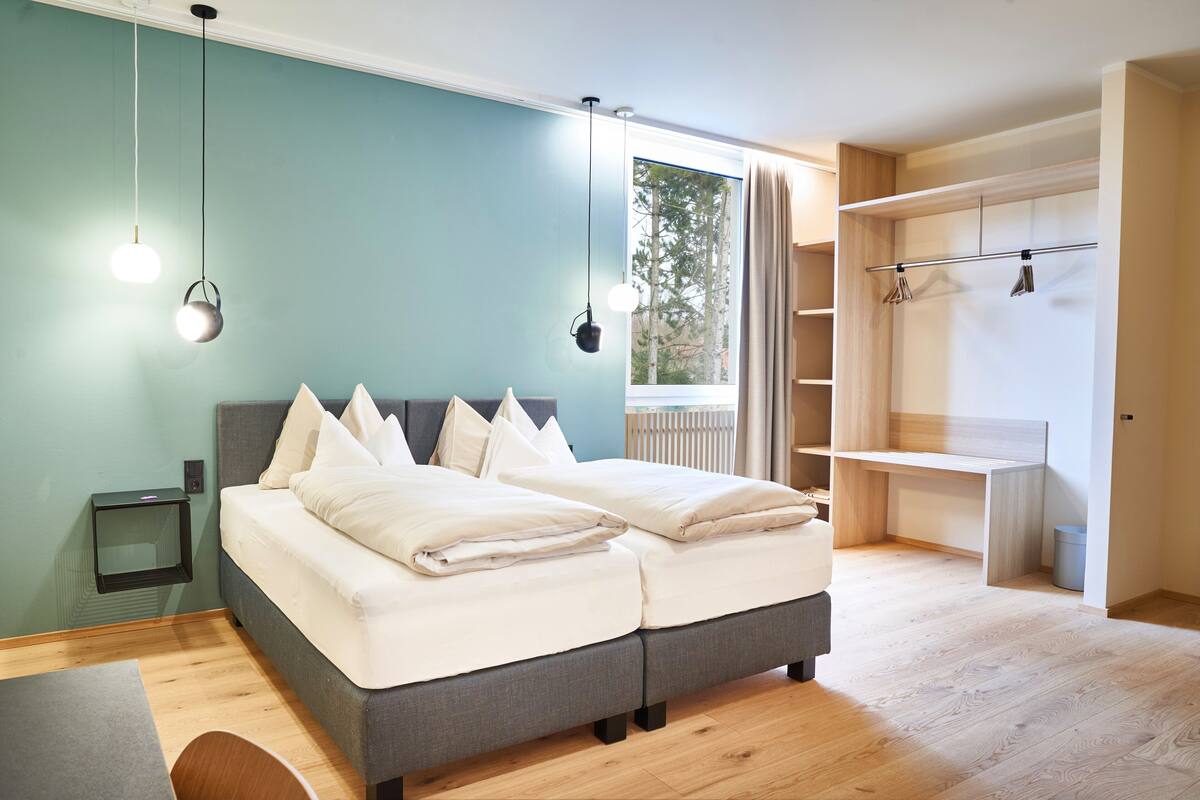 A modern bedroom with a grey bed, white bedding, a small bedside table, and a wardrobe with a light wood finish.