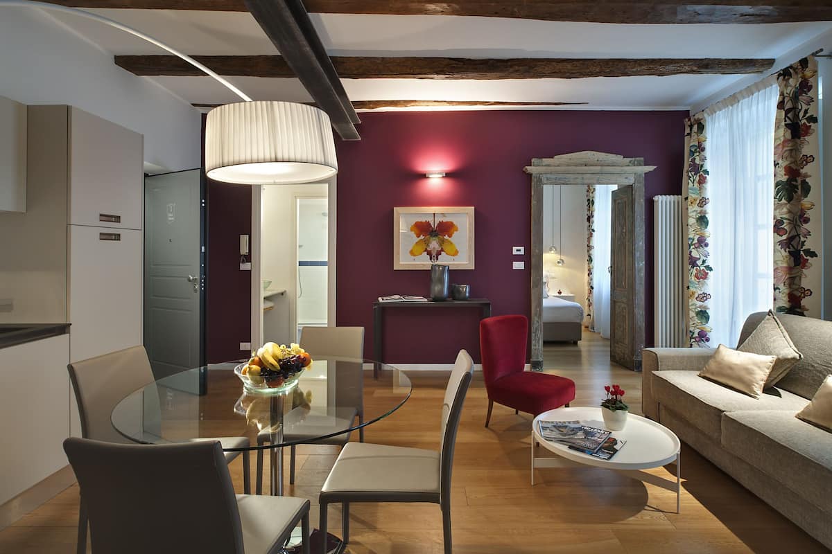 Un salon moderne avec une table à manger en verre, des chaises rouges, un canapé beige et un fauteuil rouge. Une grande fenêtre est agrémentée de rideaux à motifs floraux, et le plafond est orné de poutres apparentes.