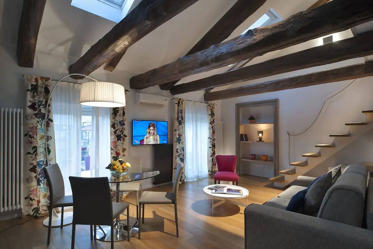 Un salon moderne avec des poutres apparentes, un coin repas doté d’une table en verre, un fauteuil rouge et un escalier menant à un autre niveau.