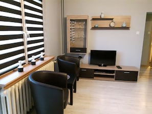 Una sala de estar moderna con televisor, sillas negras, una mesa de centro de madera y una estantería con objetos decorativos.