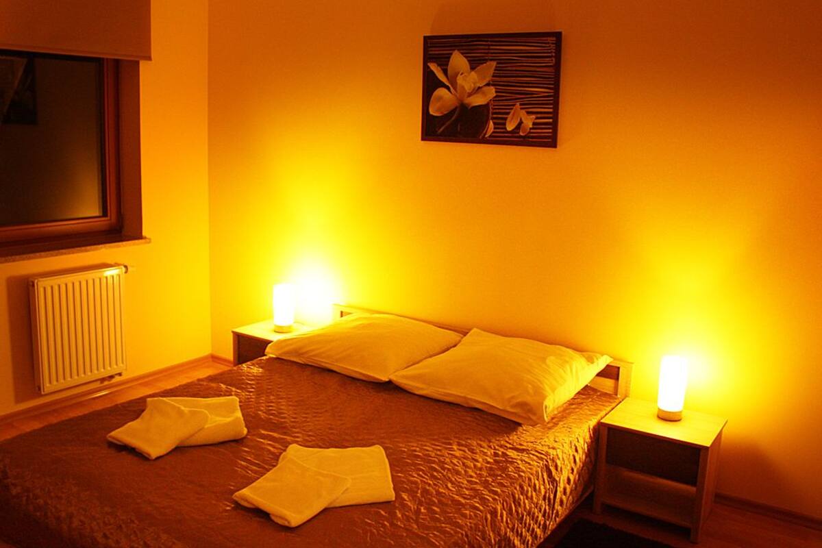 A hotel room with a bed, two bedside lamps, a window, a radiator, and a wall art piece.