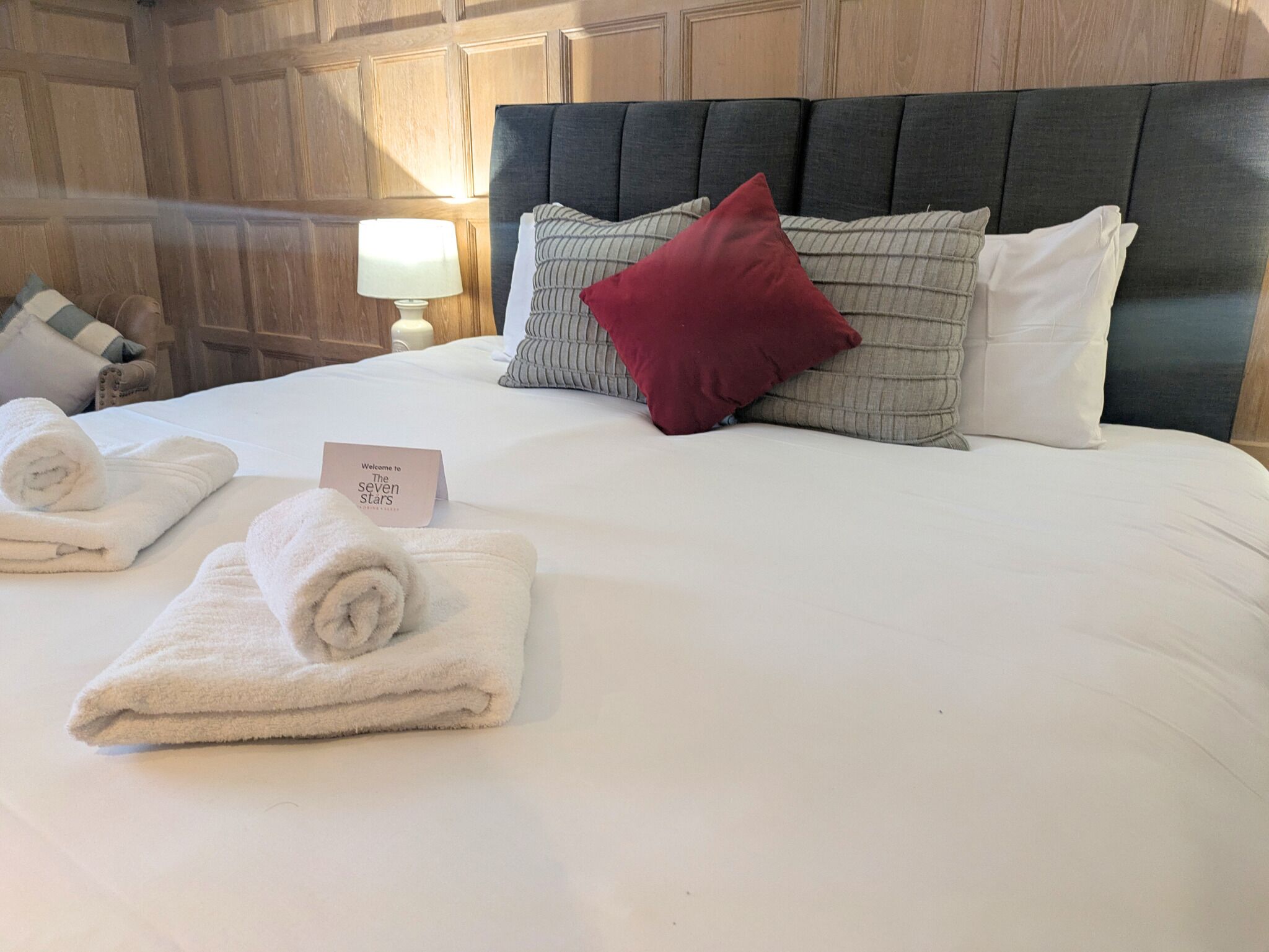 Una cama cuidadosamente tendida con sábanas blancas, un almohadón rojo y un letrero que dice "Welcome to The Seven Stars".