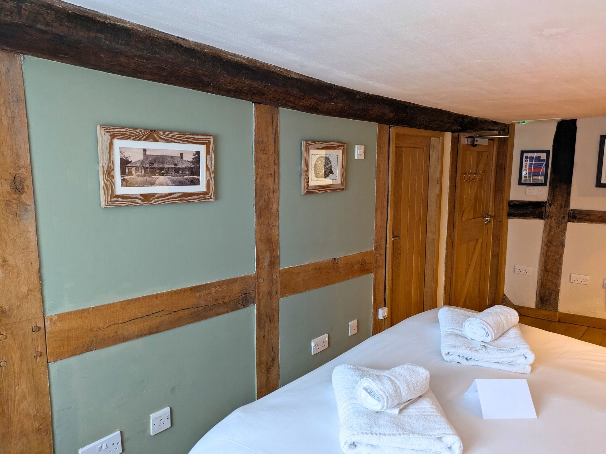 A hotel room with a bed, two framed pictures on the wall, a wooden door, and a wall-mounted thermostat.