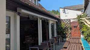 Une terrasse extérieure avec des tables et des chaises en bois, un espace salon couvert et une vue sur la rue.