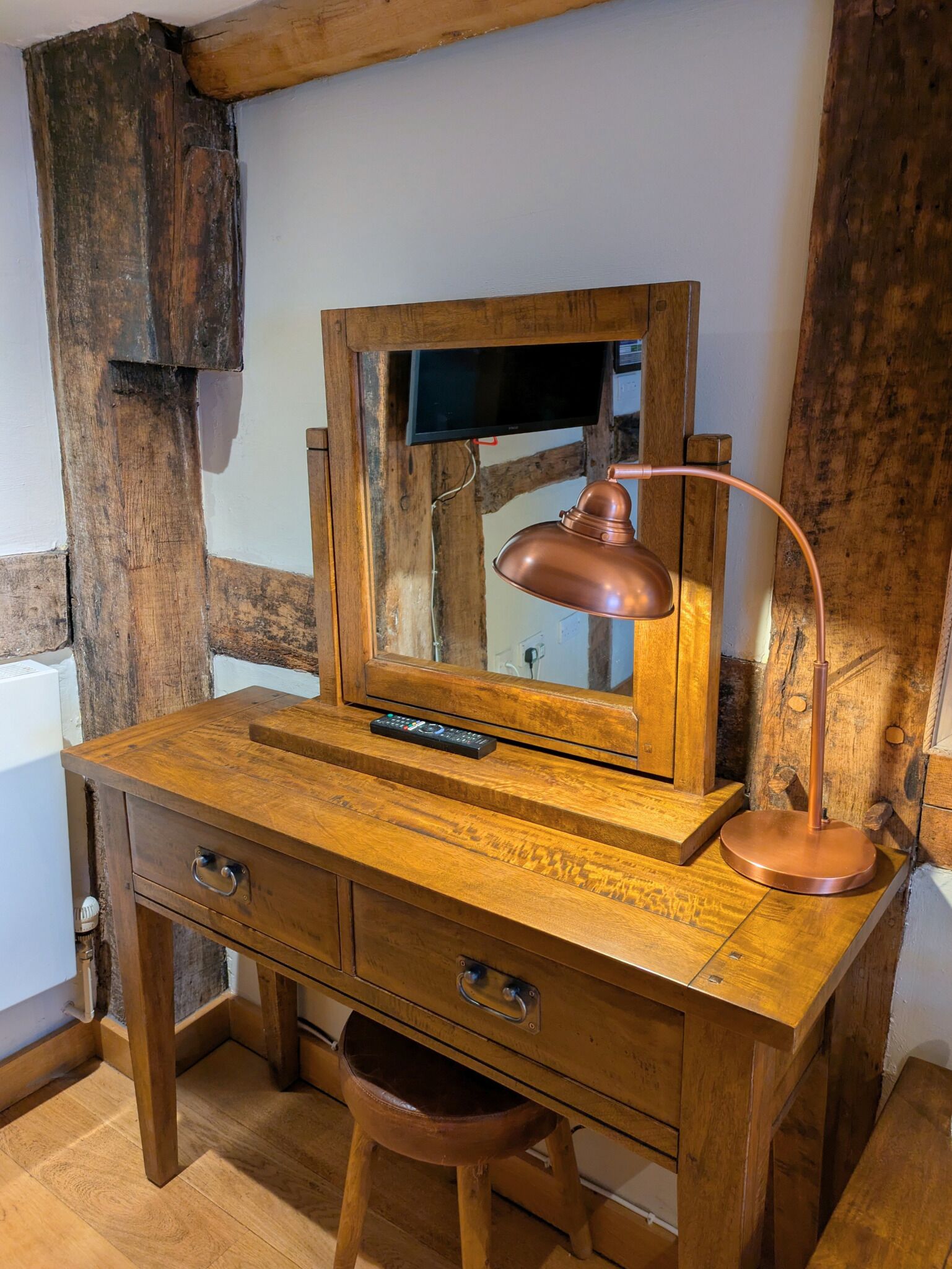 Uma escrivaninha de madeira com um espelho, uma luminária de mesa de cobre e um controle remoto.