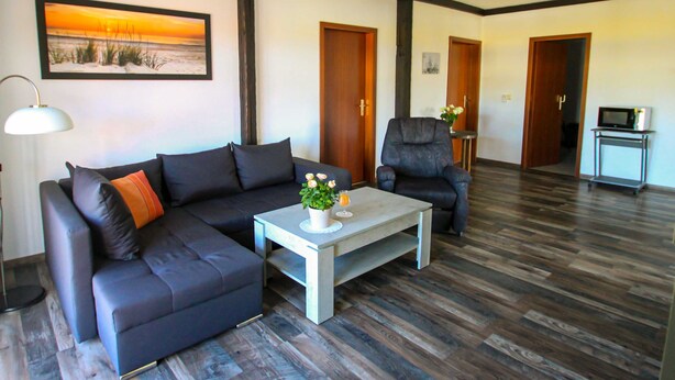 A modern living room with a dark-colored sofa, a matching armchair, a coffee table with a vase and a glass of orange juice, and a painting of a sunset on the wall.