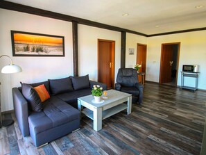 A modern living room with a dark-colored sofa, a matching armchair, a coffee table with a vase and a glass of orange juice, and a painting of a sunset on the wall.