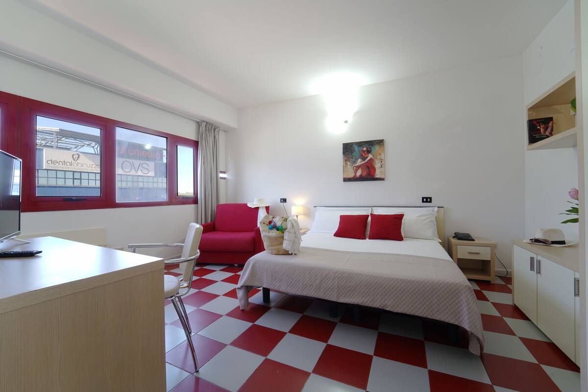 A hotel room with a checkered floor, a bed with red pillows, a desk with a computer, and a window with a view of a building.