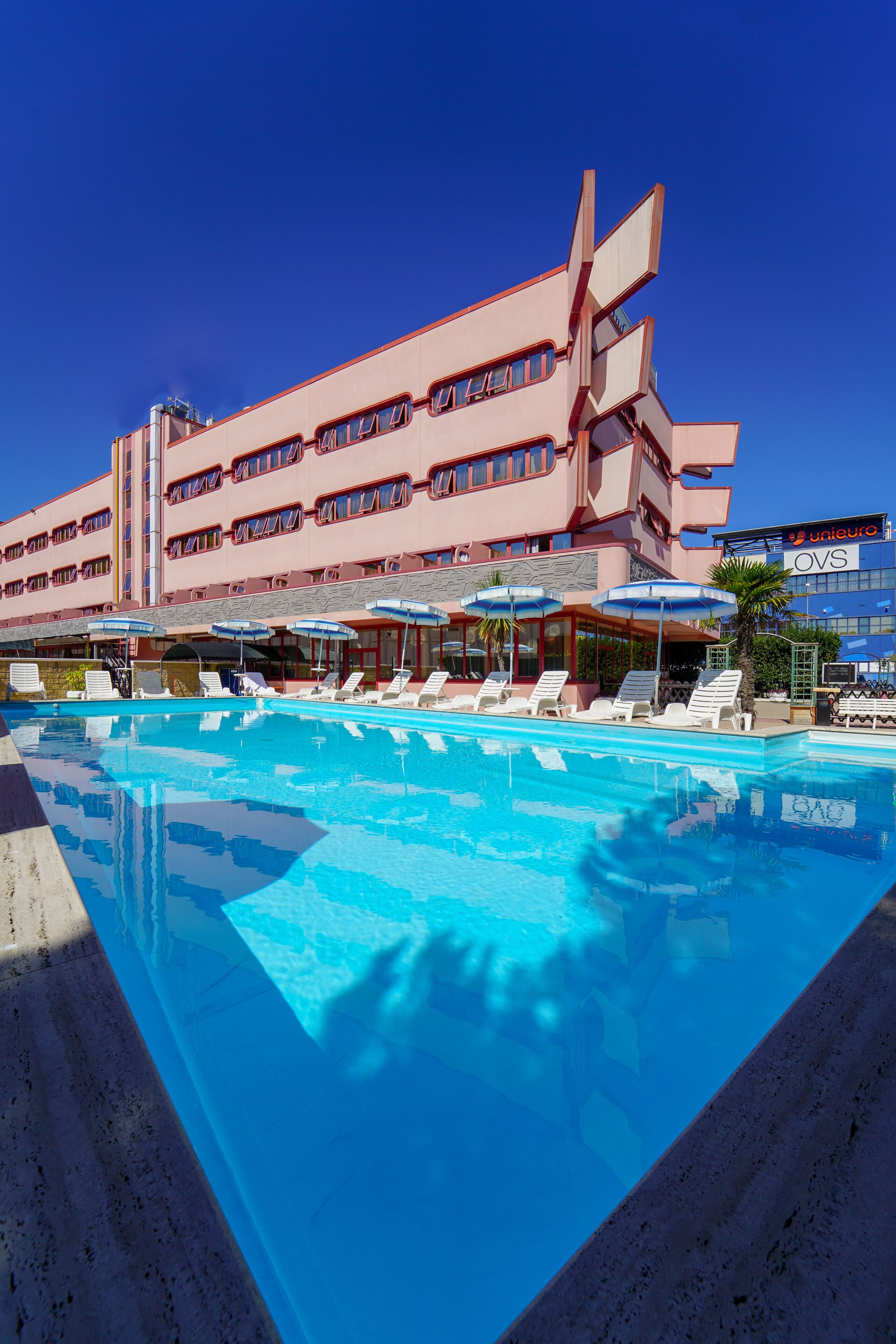 A hotel with a swimming pool, sun loungers, umbrellas, and a distinctive pink building.