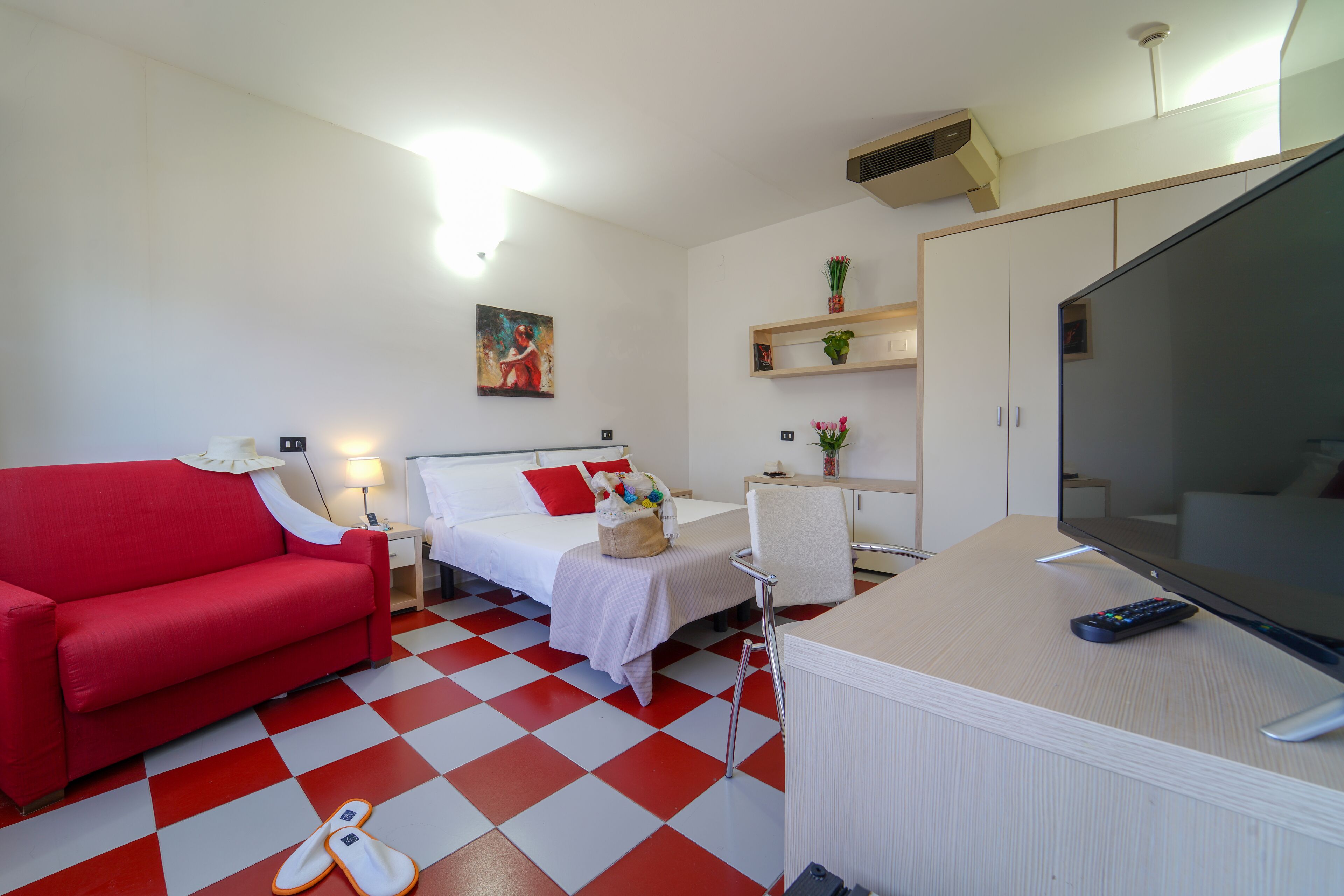 A hotel room with a red sofa, a bed with white bedding, a desk with a computer and a TV, and a checkered floor.