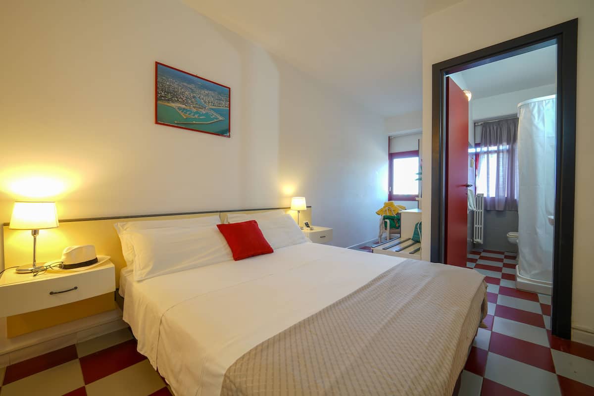 A hotel room with a bed, nightstands, a lamp, a hat, a red pillow, and a painting on the wall.
