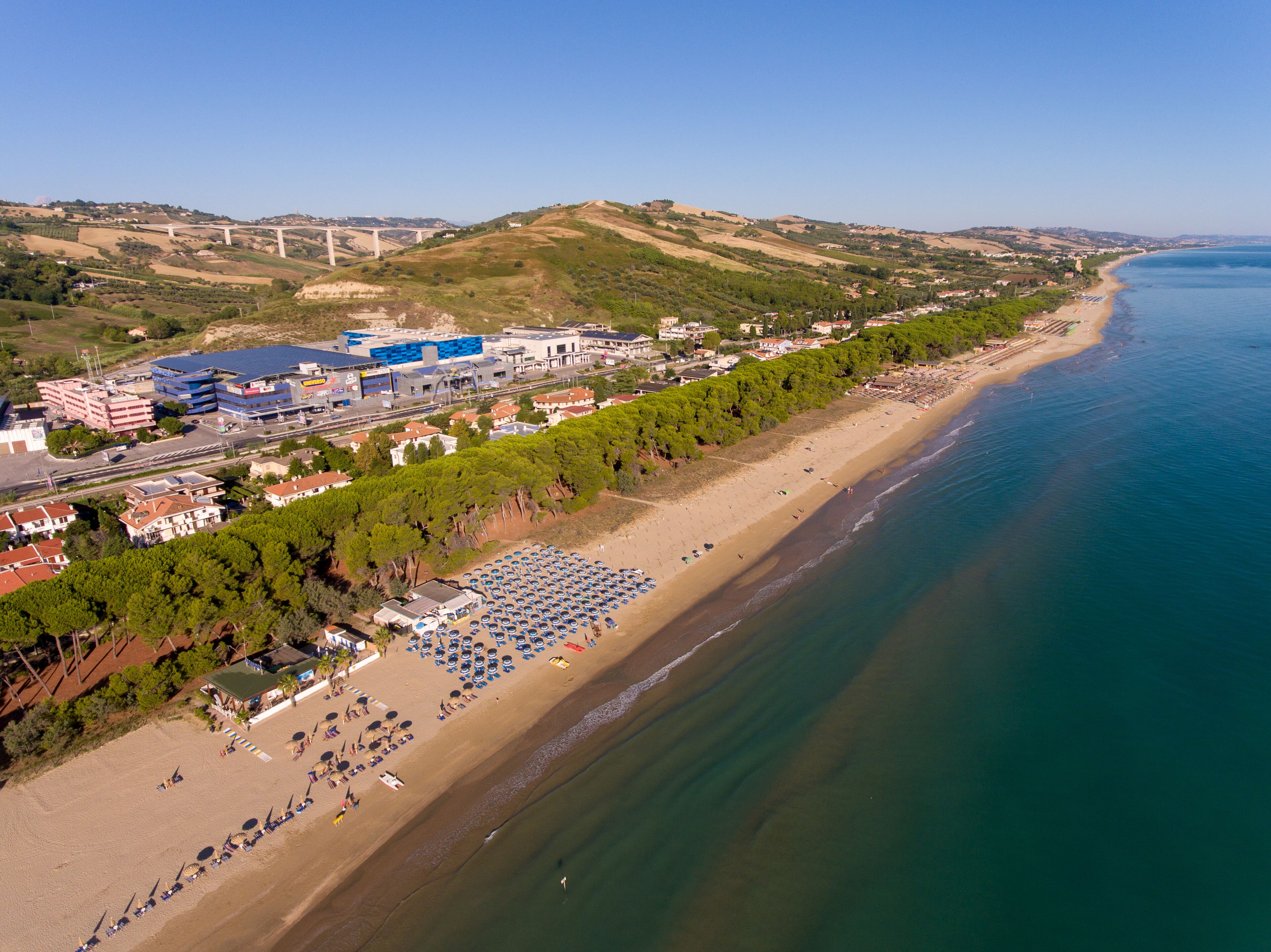 An aerial view of a coastal area with a beach, a large hotel with multiple floors, and a bridge in the background.