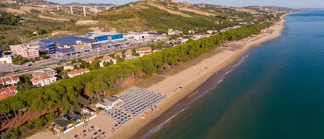 An aerial view of a coastal area with a beach, a large hotel with multiple floors, and a bridge in the background.