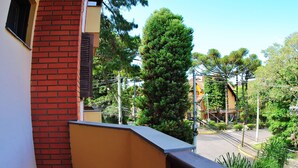 A balcony with a red brick wall and a yellow railing overlooking a street with trees and buildings. - Apartamento Serra - Gramado (Gramado)