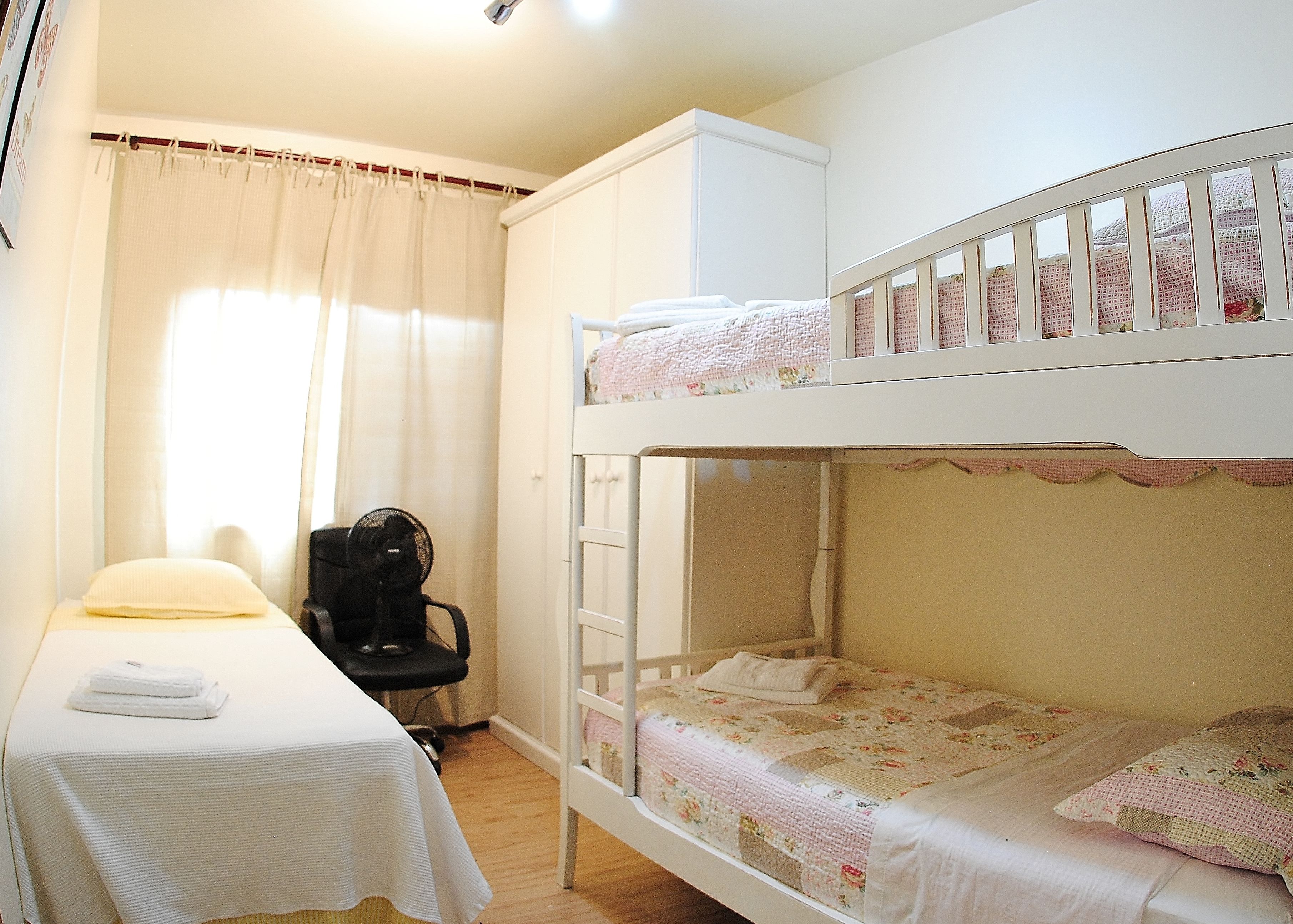 Une chambre avec un lit superposé, un bureau et une chaise, un lit recouvert d’une courtepointe à motifs floraux et une fenêtre avec des rideaux.