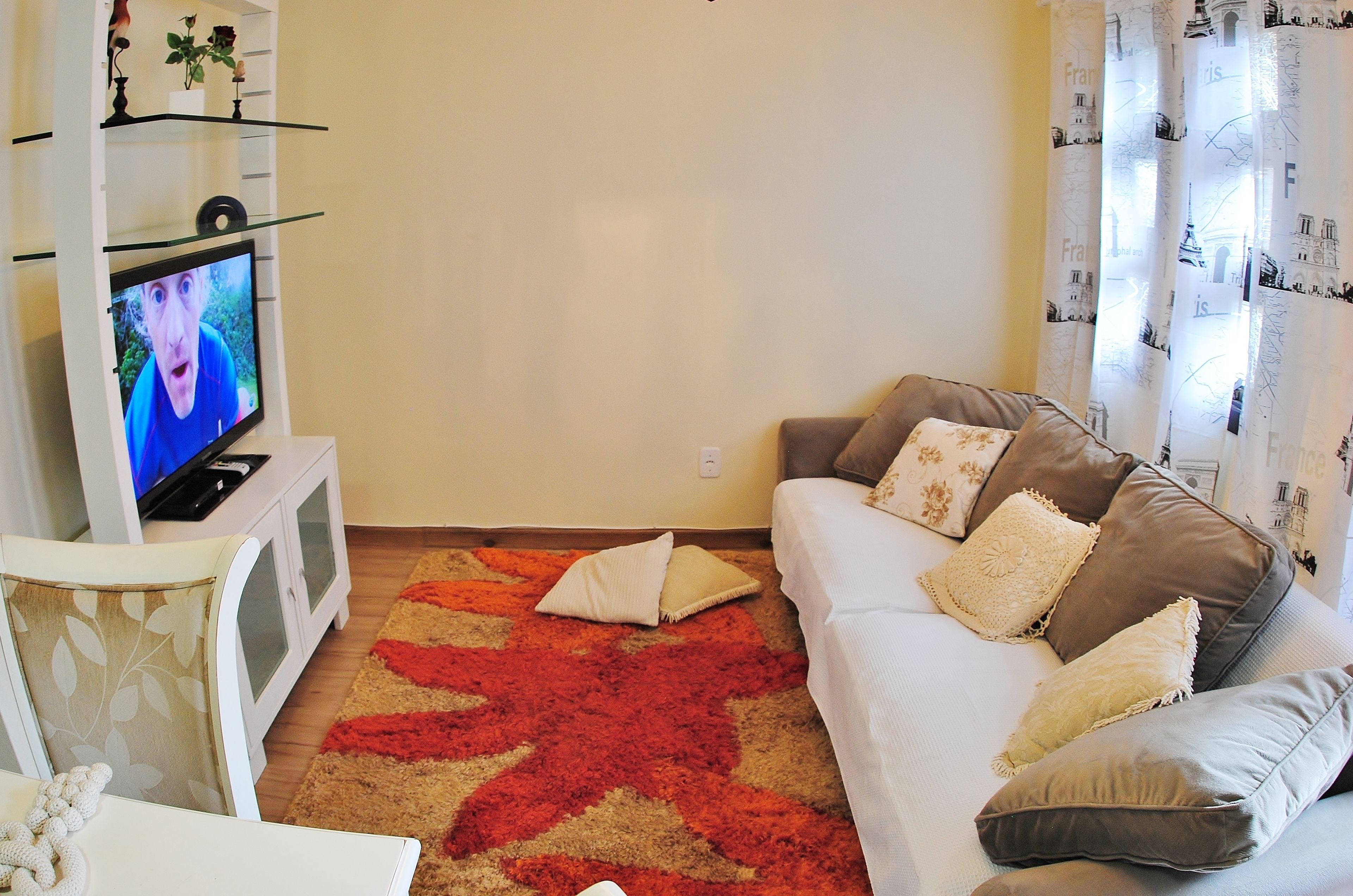 Un salón con un televisor sobre un mueble blanco, un sofá marrón con cojines y una alfombra roja estampada.