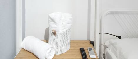 Habitación de hotel ordenada con una mesita de noche de madera, una toalla blanca, una cesta y un control remoto.