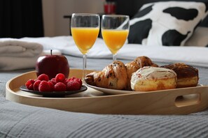 Un plateau de petit-déjeuner avec des viennoiseries, une pomme rouge, des framboises et deux verres de jus d’orange, posé sur un lit avec un oreiller noir et blanc.
