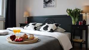 Un lit bien fait, agrémenté d’un coussin à motifs de vaches, un plateau de petit-déjeuner comprenant une pomme, des framboises et des croissants, et un vase de tulipes posé sur une table de chevet.