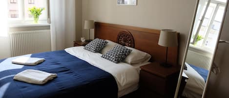 A neatly made bed with a blue and white bedspread, two pillows, and two folded towels. There is a bedside table with a lamp, a window with a view of buildings, and a framed picture on the wall.