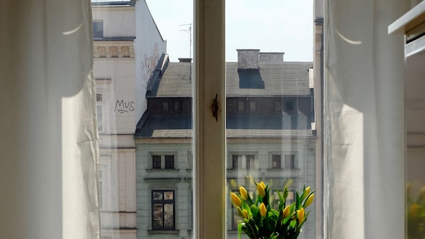 Une fenêtre avec des rideaux blancs, un vase rempli de fleurs jaunes et deux petites bougies posées sur le rebord de la fenêtre.