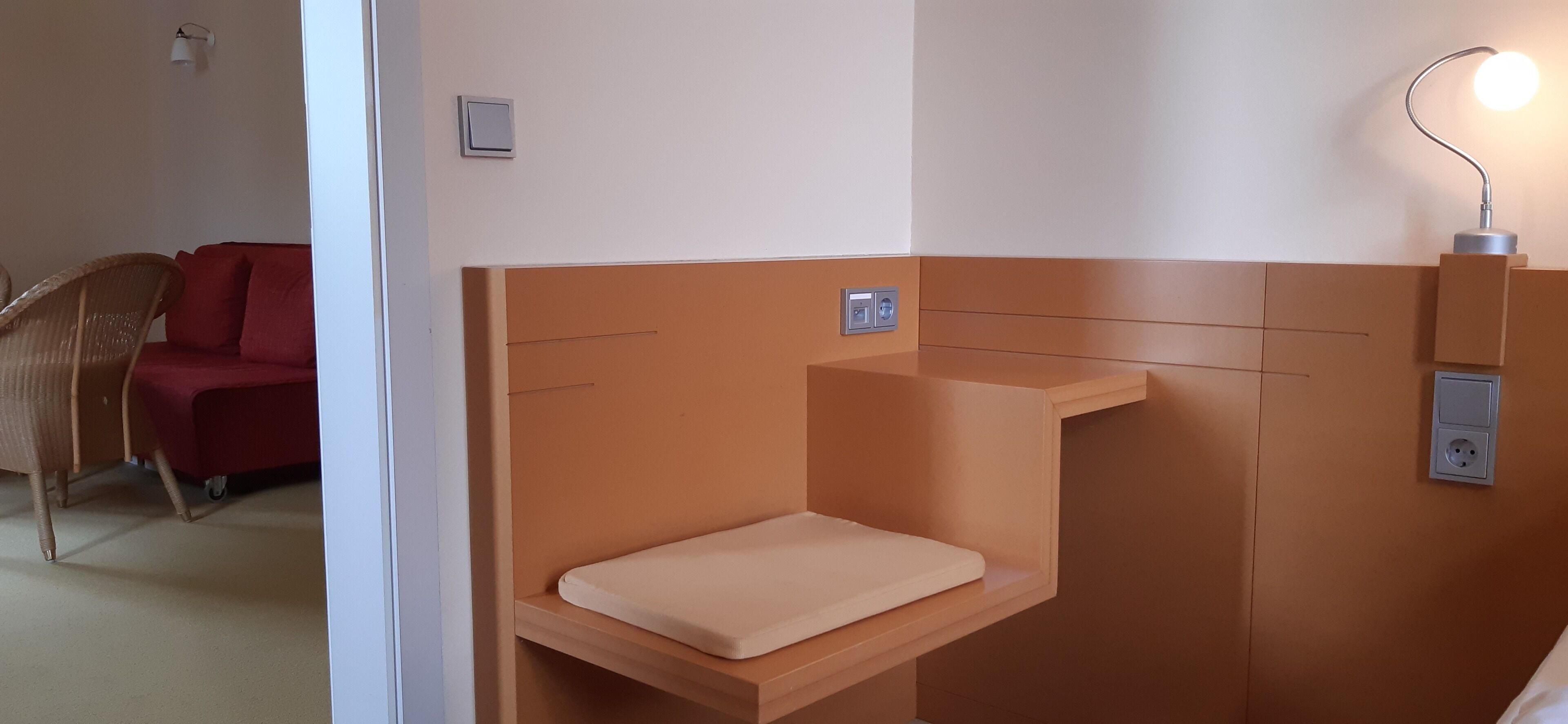 Una camera d'albergo con un letto singolo, un comodino, una lampada a parete e un divano rosso nella stanza adiacente.