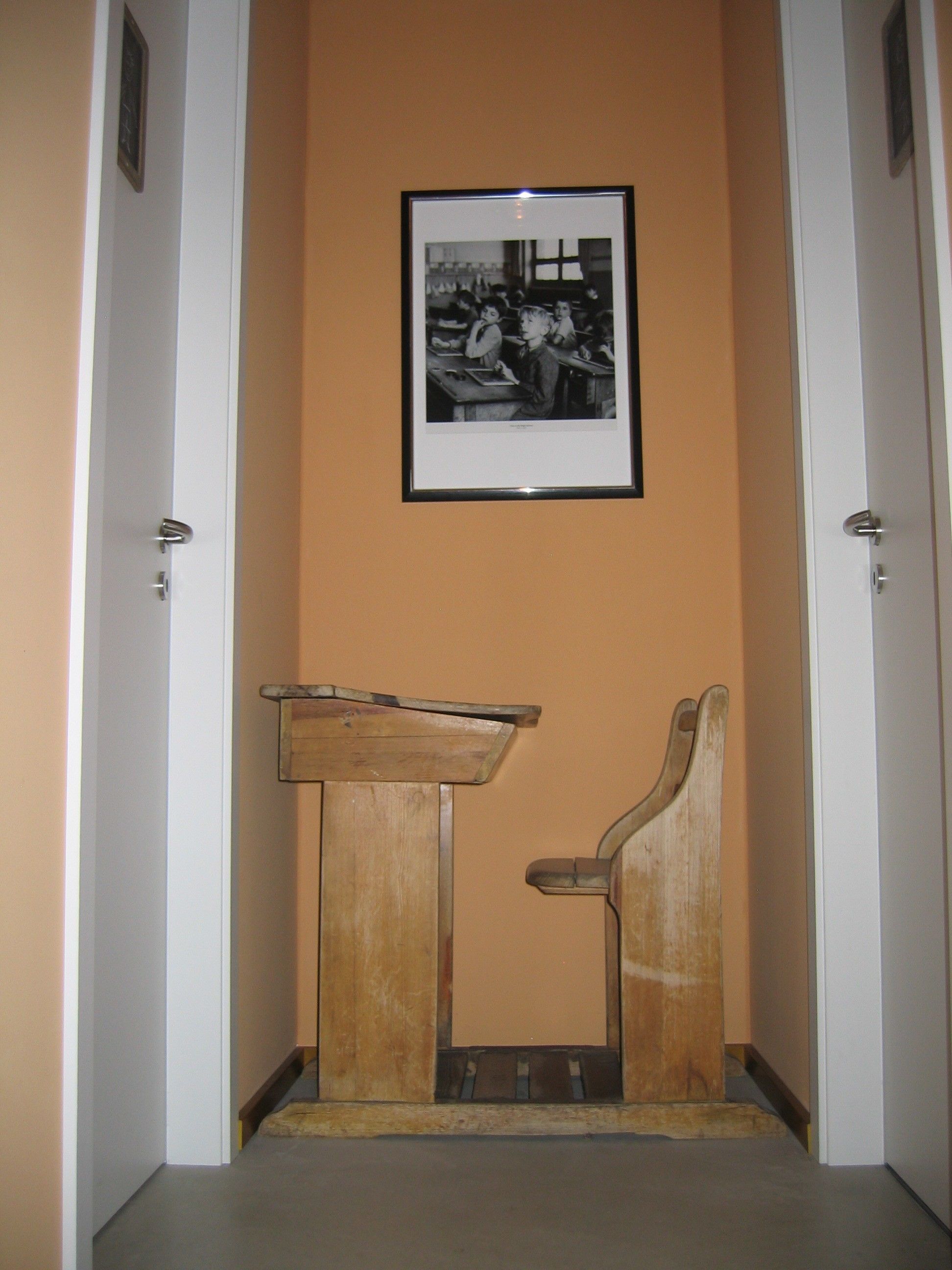 Un pasillo con un escritorio y una silla de madera, una fotografía en blanco y negro enmarcada en la pared, y puertas blancas a cada lado.