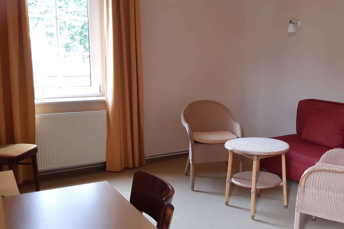 A room with a red sofa, two wicker chairs, a small round table, a radiator, and a window with curtains.
