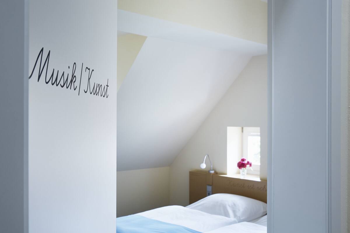 A bedroom with a sloped ceiling, a bed with white bedding, a nightstand with a lamp, and a window with flowers.