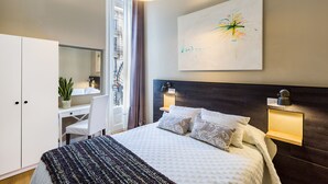 A modern hotel room with a large bed, a desk, a chair, a potted plant, a window with curtains, and a wall-mounted abstract painting.