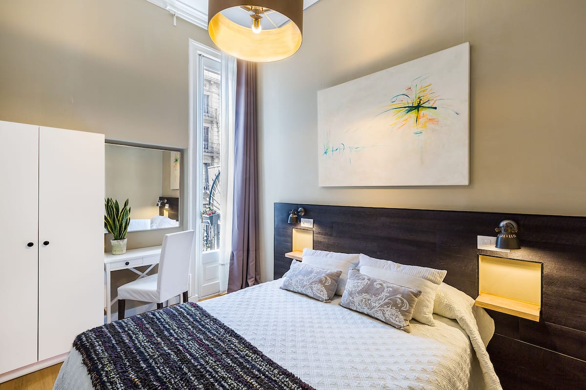 Ein modernes Hotelzimmer mit einem großen Bett, einem Schreibtisch, einem Sessel, einem Blumentopf, einem Fenster mit Vorhängen und einem an der Wand hängenden abstrakten Gemälde.